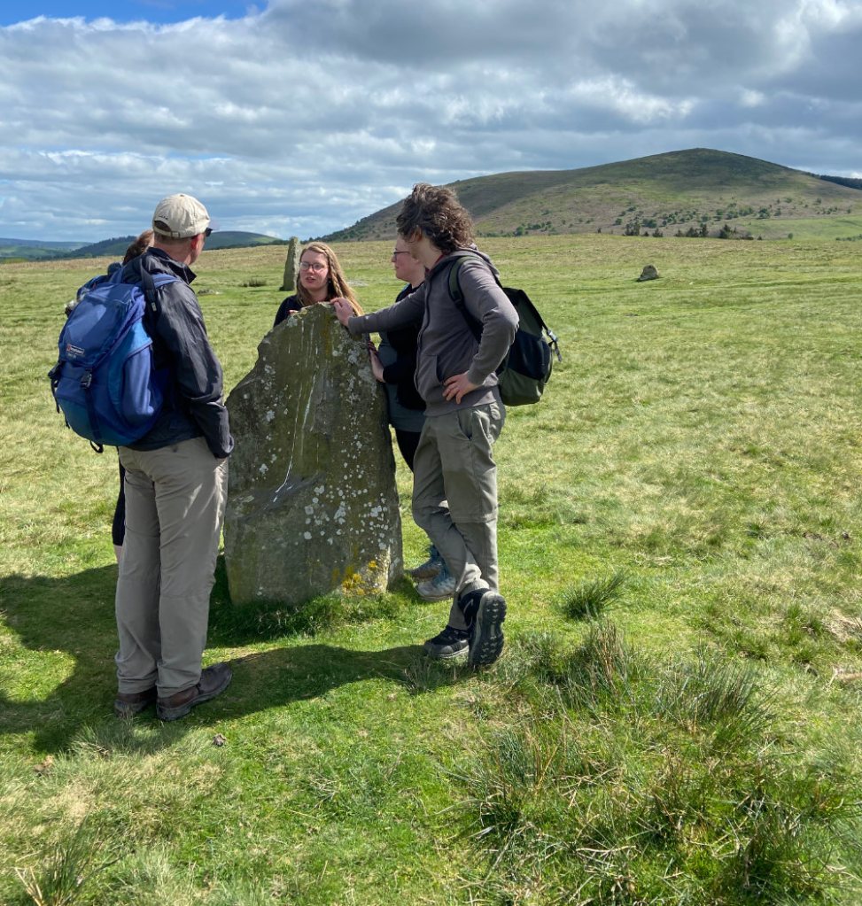 A group of walkers gather around a shoulder-height standing stone against a moorland backdrop