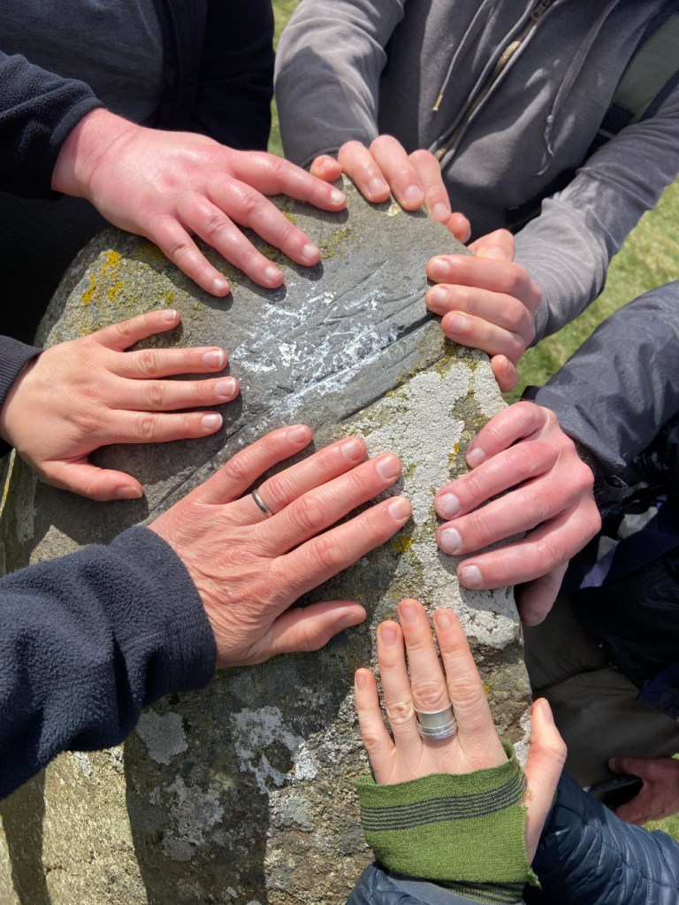 Close-up of six walkers' hands placed on a standing stone