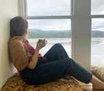A white woman with brown hair sits in front of a window with sea view, drinking a cup of tea